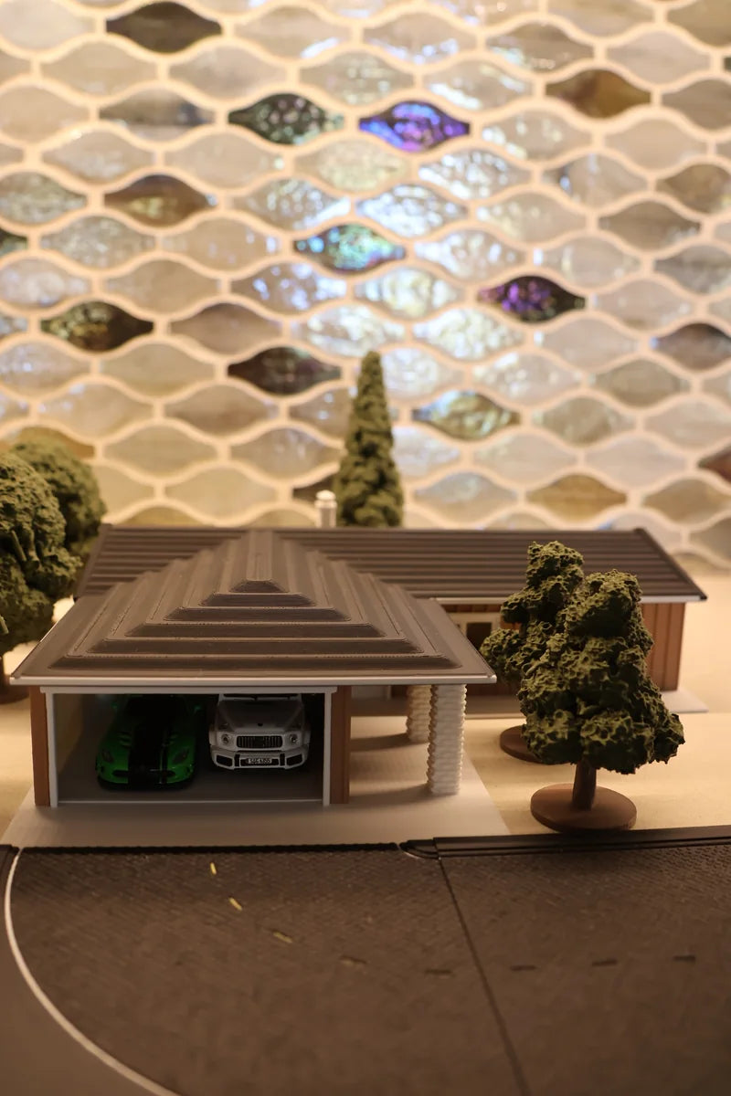 A low cream-walled printed house with a flat overhanging roof, photographed square into its open garage bay: a green car and a silver car parked inside, a round-headed tree and a conifer alongside, grey road surface in the foreground.