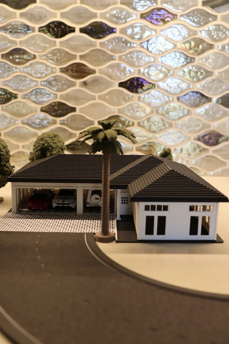 A white printed house with a dark hip roof on a corner plot: two open garage bays with cars inside, a tall palm rising past the roofline, white-framed windows along the right-hand wing, and a road tile curving past the front.
