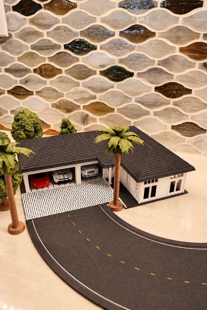 A printed single-storey house from high above: the dark shingled hip roof open to the lens, two garage bays with cars inside below it, a palm alongside, and the grey road tile curving right across the frame with its far edge still in shot.