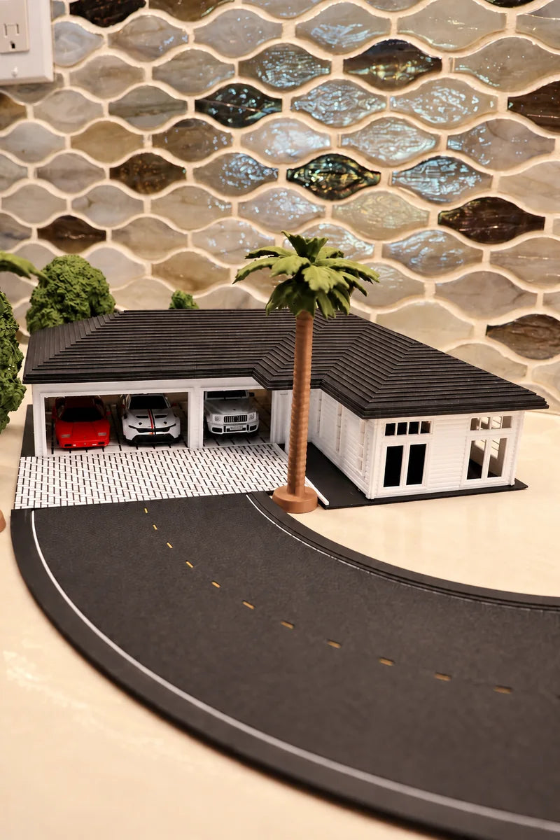 The same house from shelf height, turned so front and side are both in view: the hip roof foreshortened, two open bays with a red car and a silver car inside, a palm beside them, and the road tile running out of the bottom of the frame.