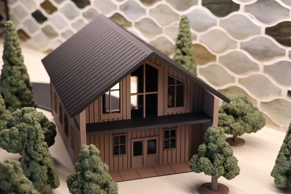 A two-storey printed house with a steep dark standing-seam metal roof over vertical board siding, tall windows in the gable end, ringed by conifers and round-headed trees.