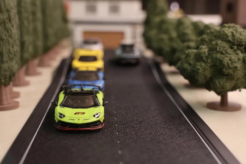 Four cars nose to tail coming down a printed road between rows of trees, a lime green Lamborghini leading with blue, yellow and dark cars behind it, and a white house with its garage bay open at the end of the street.