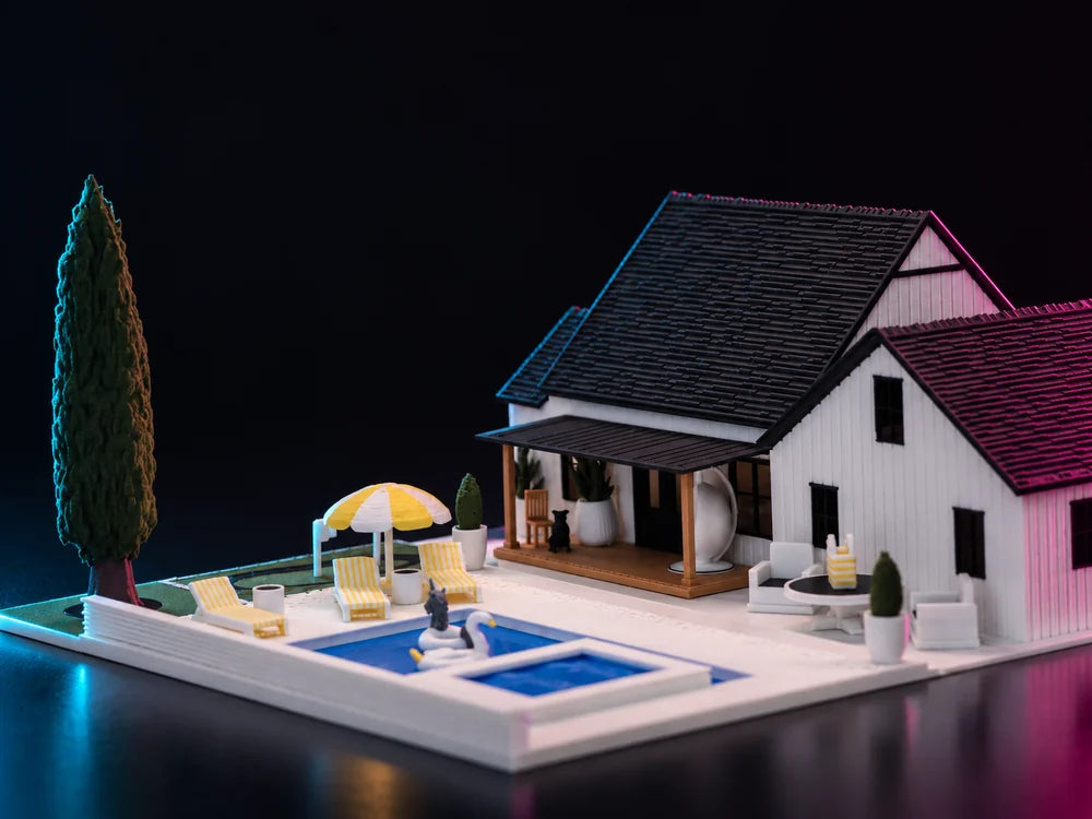 A printed white house with a steep dark shingled roof standing on a black plinth at night, a blue pool and a yellow parasol on the deck in front of it and a tall dark cypress at the left.