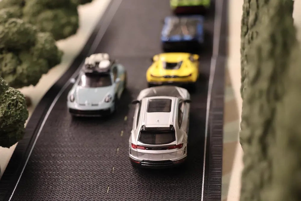 Looking down on two lanes of a printed road: a silver Lamborghini in the near lane, a pale blue Porsche passing the other way, and yellow, blue and green cars strung out ahead between the trees.