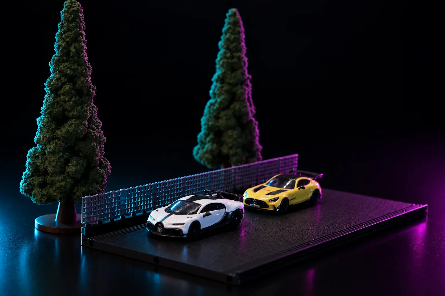 A printed display base in charcoal lit in blue and magenta, an open square-mesh fence running along the back edge with two conifers standing behind it and two 1:64 cars parked side by side on the deck.