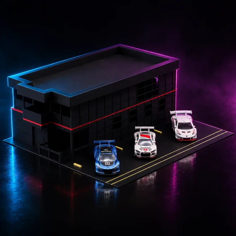 A printed two-storey racing paddock in black with blue and magenta neon edge lighting, three GT cars parked on the pit lane in front of it.