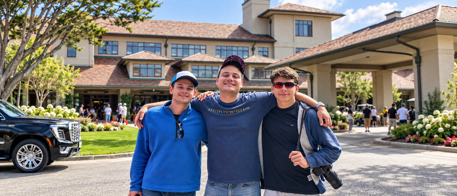 Three young men standing arm in arm in bright daylight outside
                a large shingled clubhouse, a black SUV parked at the left and
                white hydrangeas along the drive at the right.